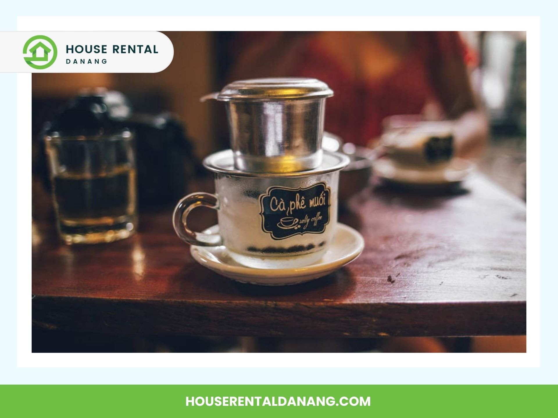 A Vietnamese drip coffee sits on a wooden table with a glass of water alongside. A person is partially visible in the background. The image border features a "House Rental Danang" logo and website, hinting at nearby adventures like the Best Places for Shopping in Da Nang.
