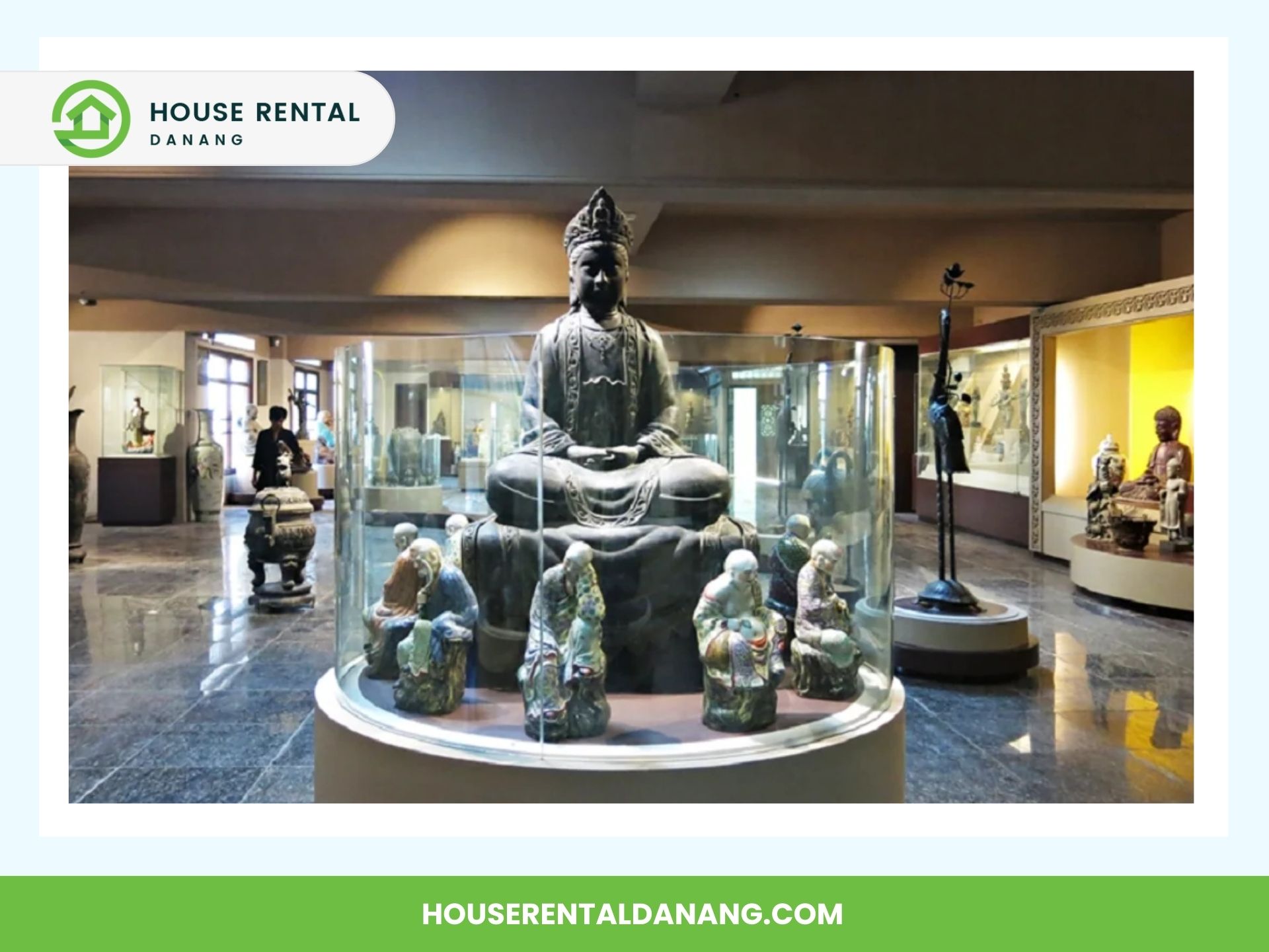 A museum exhibit at the Museum of Cham Sculpture in Da Nang features a large statue of a seated figure surrounded by smaller statues, all encased in a glass display. The room boasts several other artifacts and statues meticulously placed in glass cases.