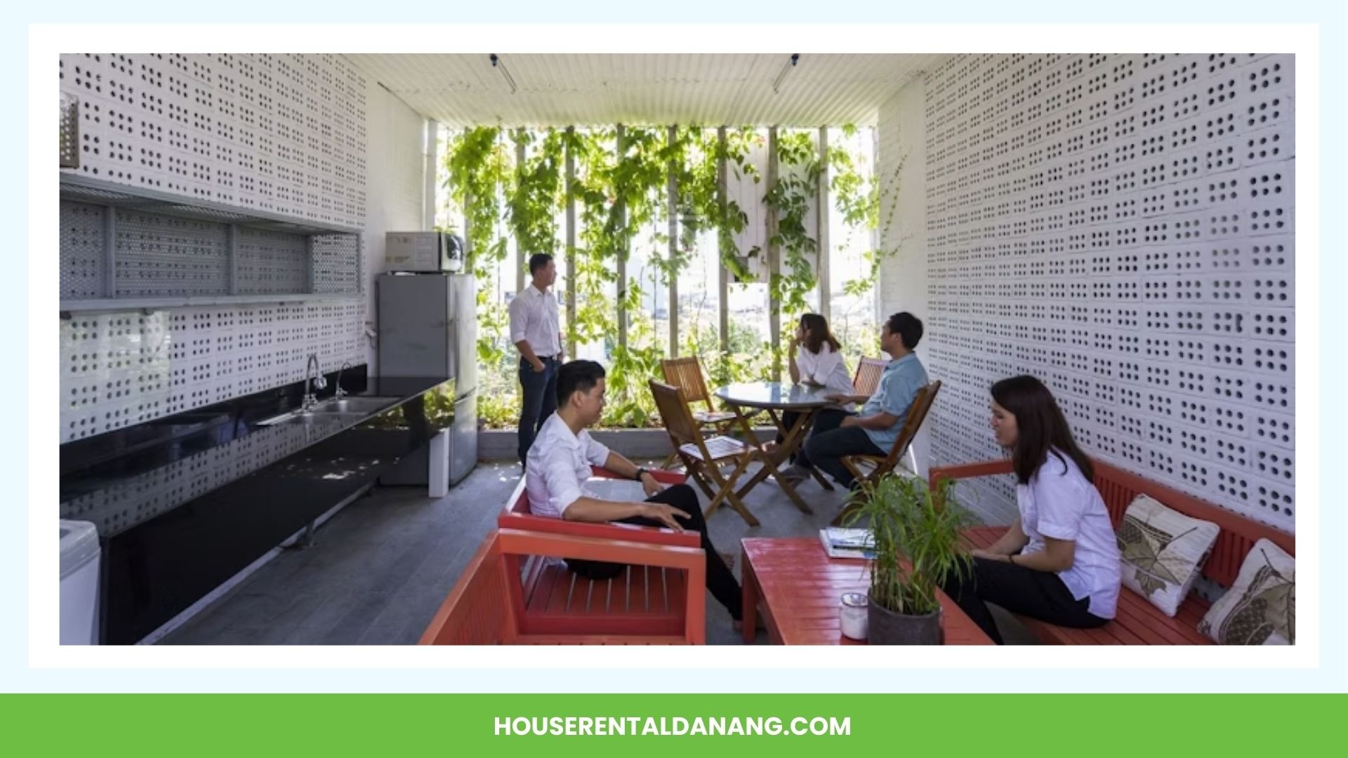 Modern indoor patio with white perforated walls, green vine plants, assorted seating, tables, and several people sitting and conversing. Ideal for those exploring coworking spaces in Da Nang. "HOUSERENTALDANANG.COM" is displayed at the bottom.