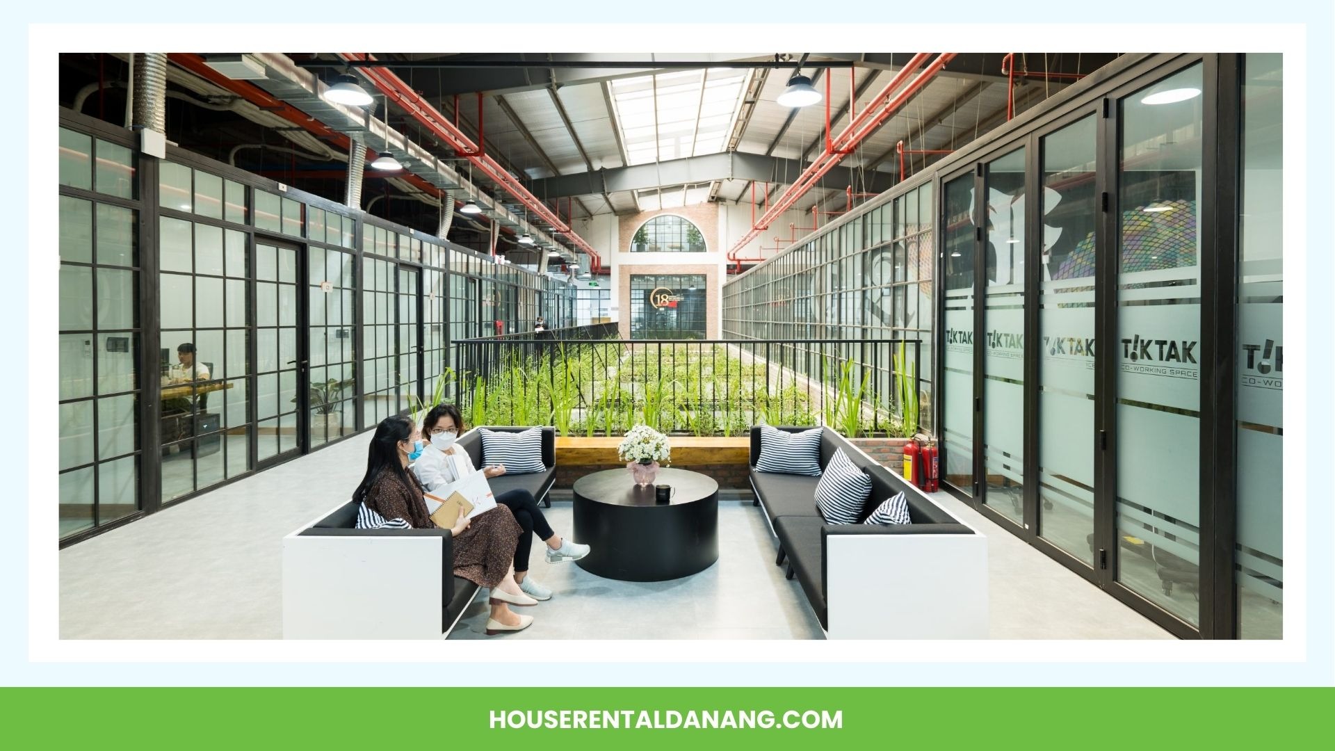 Three people wearing masks sit on modern outdoor furniture in a spacious atrium, reminiscent of the coworking spaces in Da Nang. The area boasts large glass windows, a garden in the center, and an industrial ceiling.