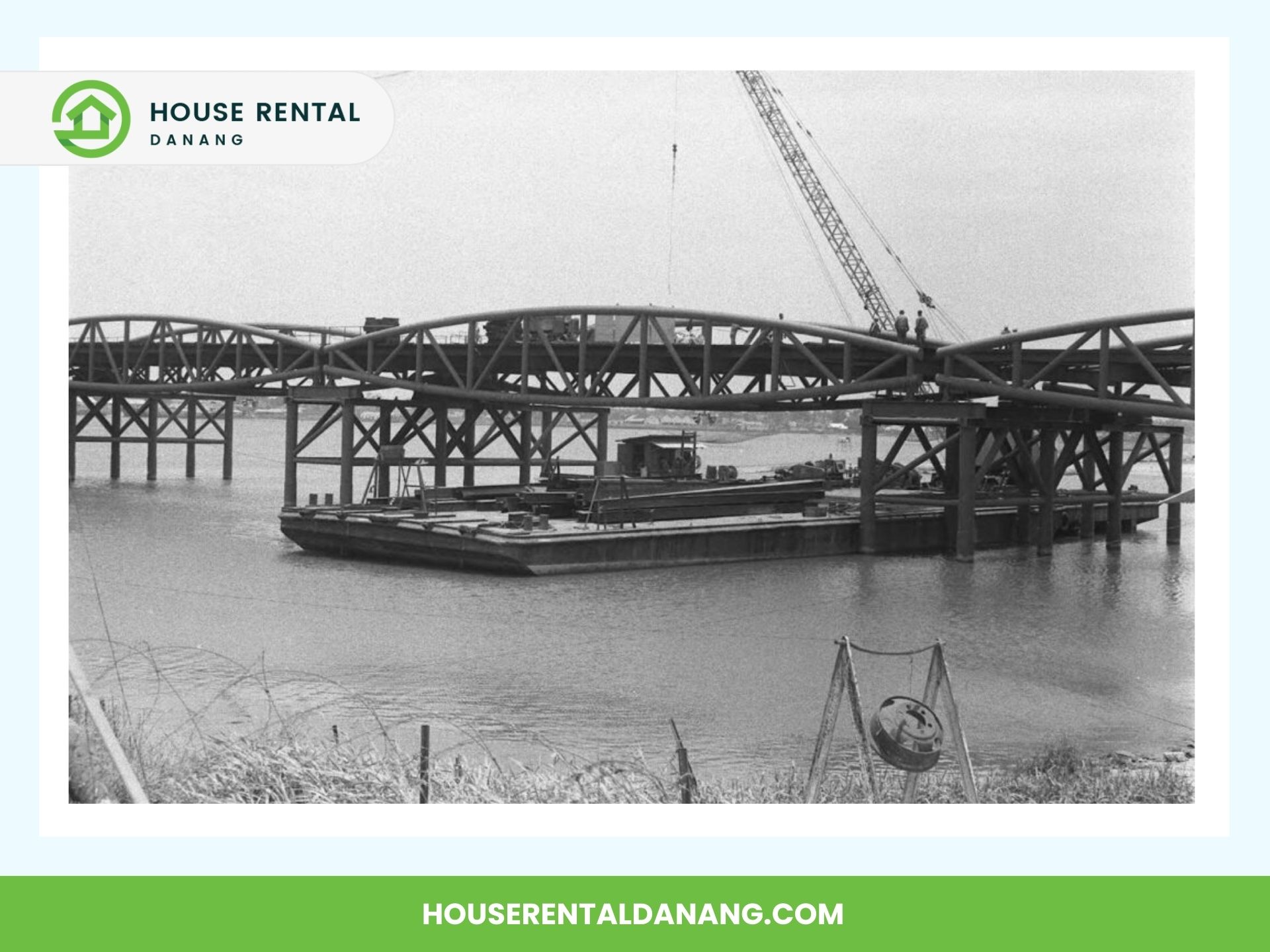 Black and white photo of Nguyen Van Troi Bridge under construction over the Han River in Da Nang, with workers and a crane on the bridge, and a barge in the water below. Text reads "House Rental Danang" and "HOUSERENTALDANANG.COM".