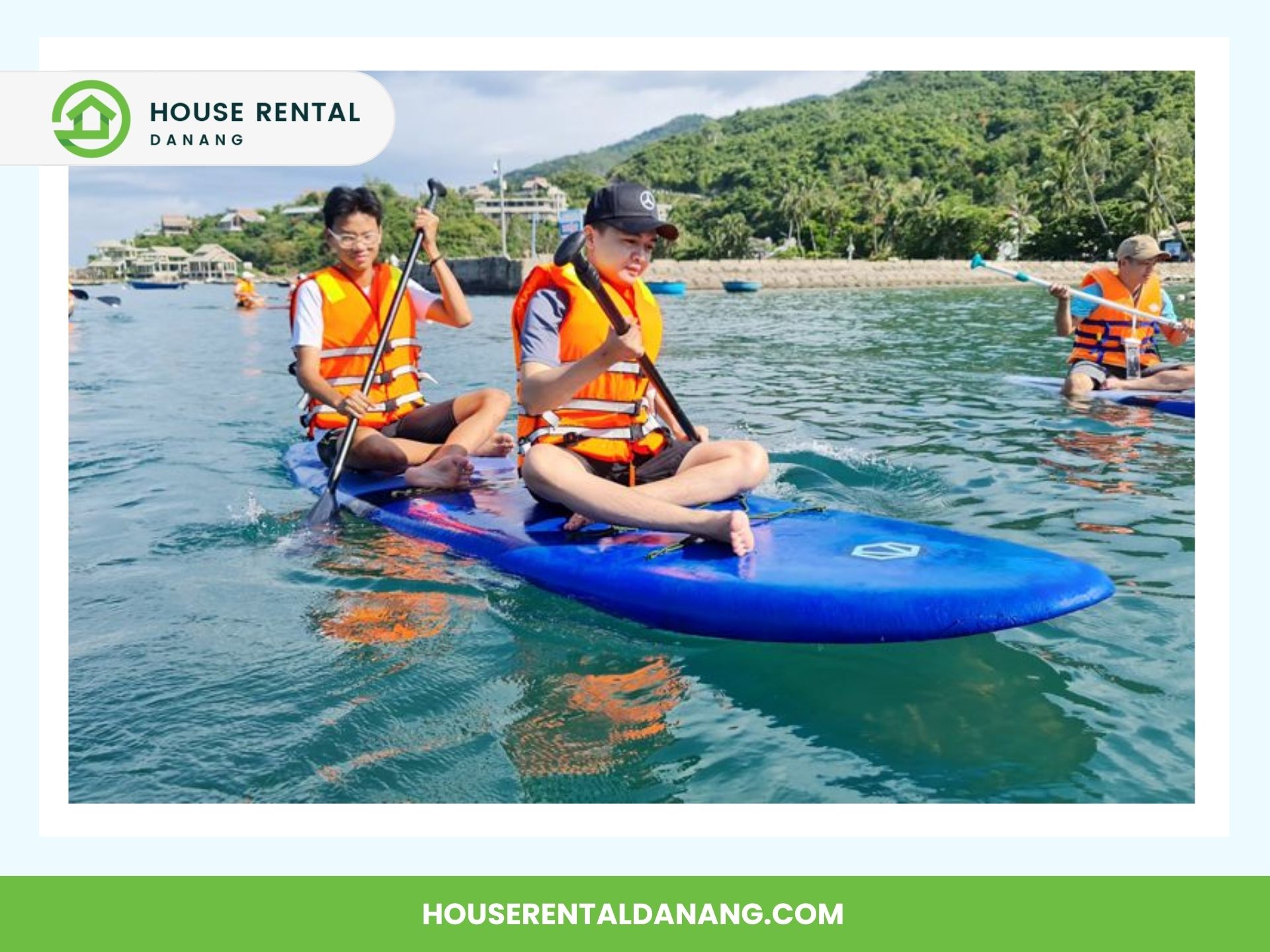 Two people wearing life vests paddle on a blue inflatable kayak in calm water, surrounded by greenery and residential buildings in the background. Discover the joy of kayaking at Cham Island, one of many reasons travel enthusiasts can't resist this picturesque destination.