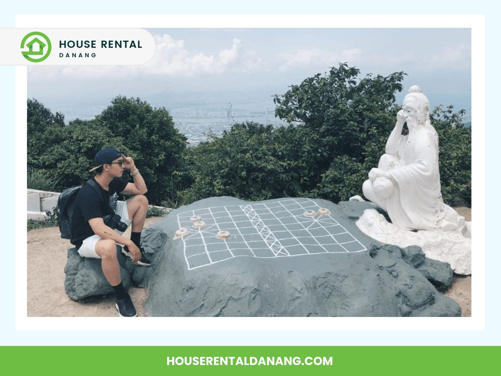 A person sits on a rock next to a statue of a seated figure, both facing a large outdoor chessboard at Ban Co Peak. Trees and cityscape are visible in the background. Text: "House Rental Danang" and website URL.