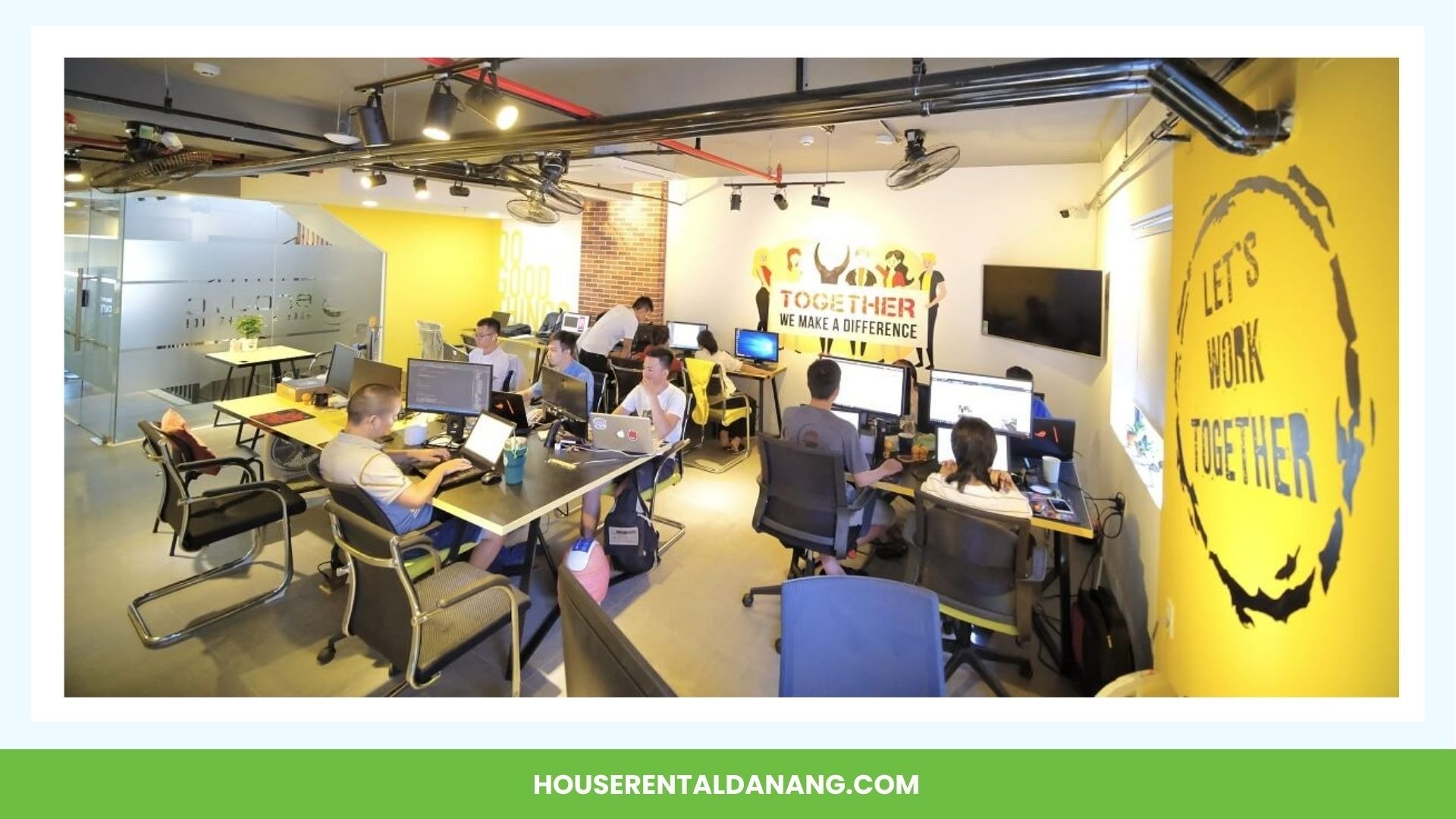 A busy office with several people working at desks with computers, surrounded by motivational posters on the yellow walls, and a green banner at the bottom with the text "HOUSERENTALDANANG.COM," showcases one of the premier coworking spaces in Da Nang.