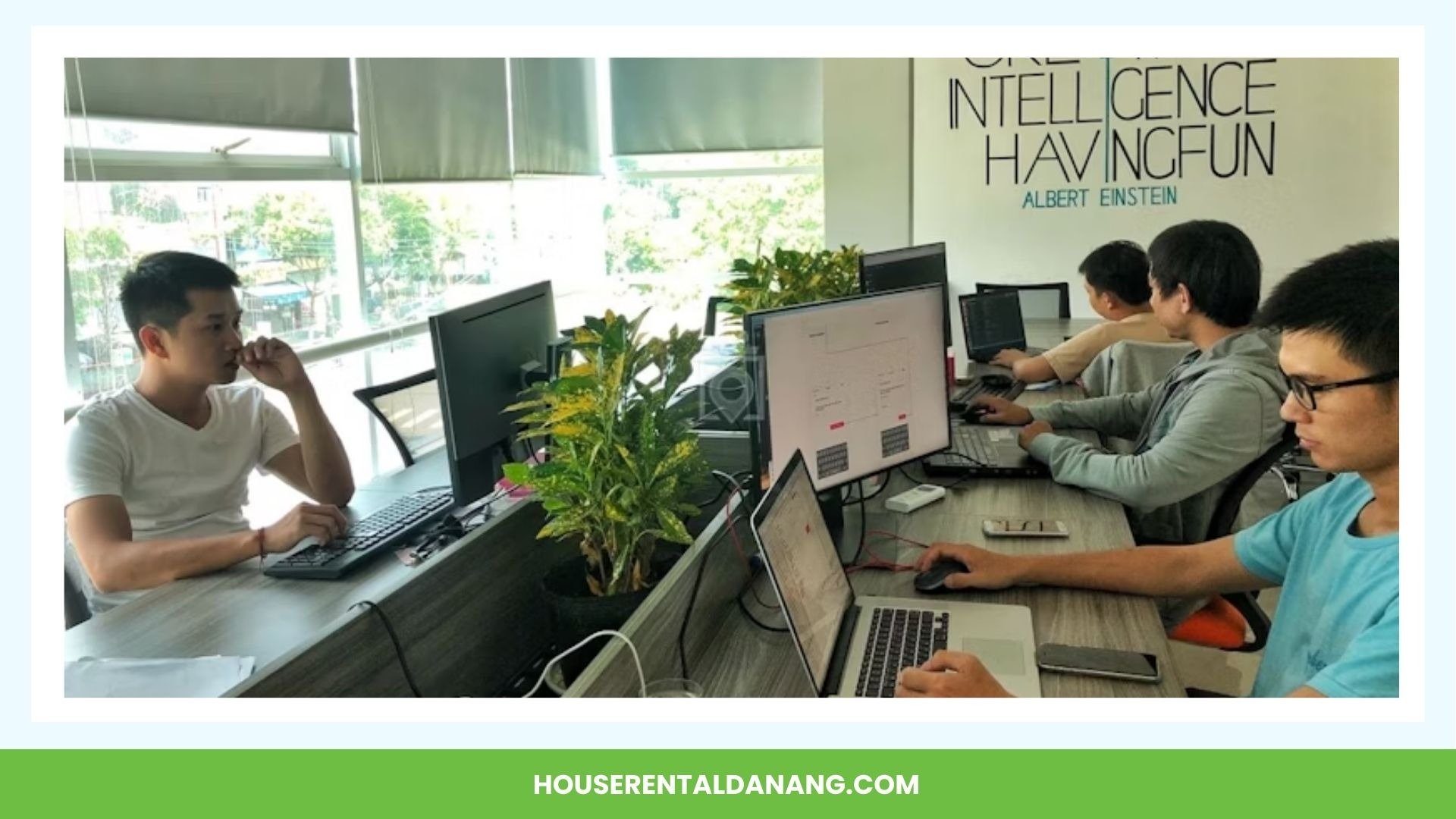 Four people are working on laptops and computers at a shared desk in an office with large windows. A quote from Albert Einstein is visible on the wall, creating an inspiring atmosphere typical of coworking spaces in Da Nang.