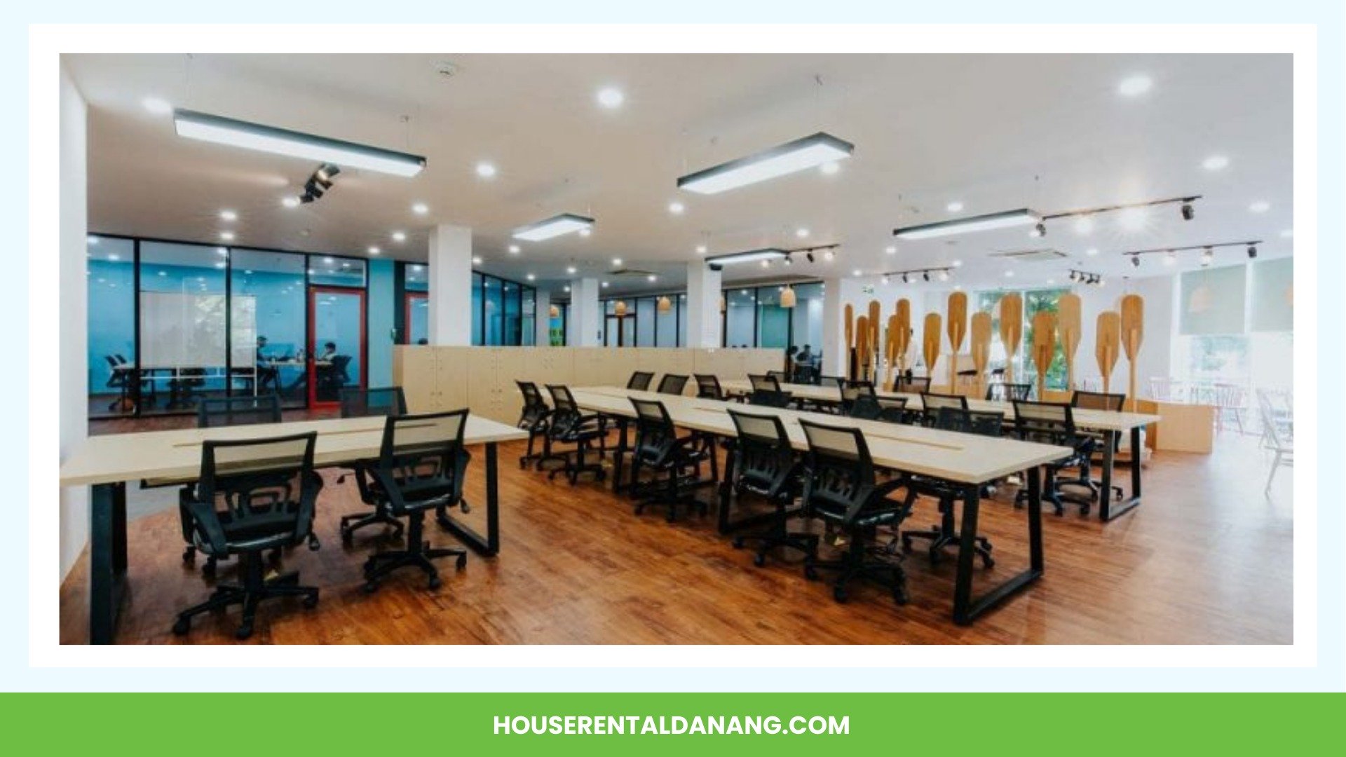 A spacious, modern office with multiple desks and chairs on a wooden floor, featuring large windows and ample lighting. The area includes partitions and a greenery backdrop, typical of the coworking spaces in Da Nang.