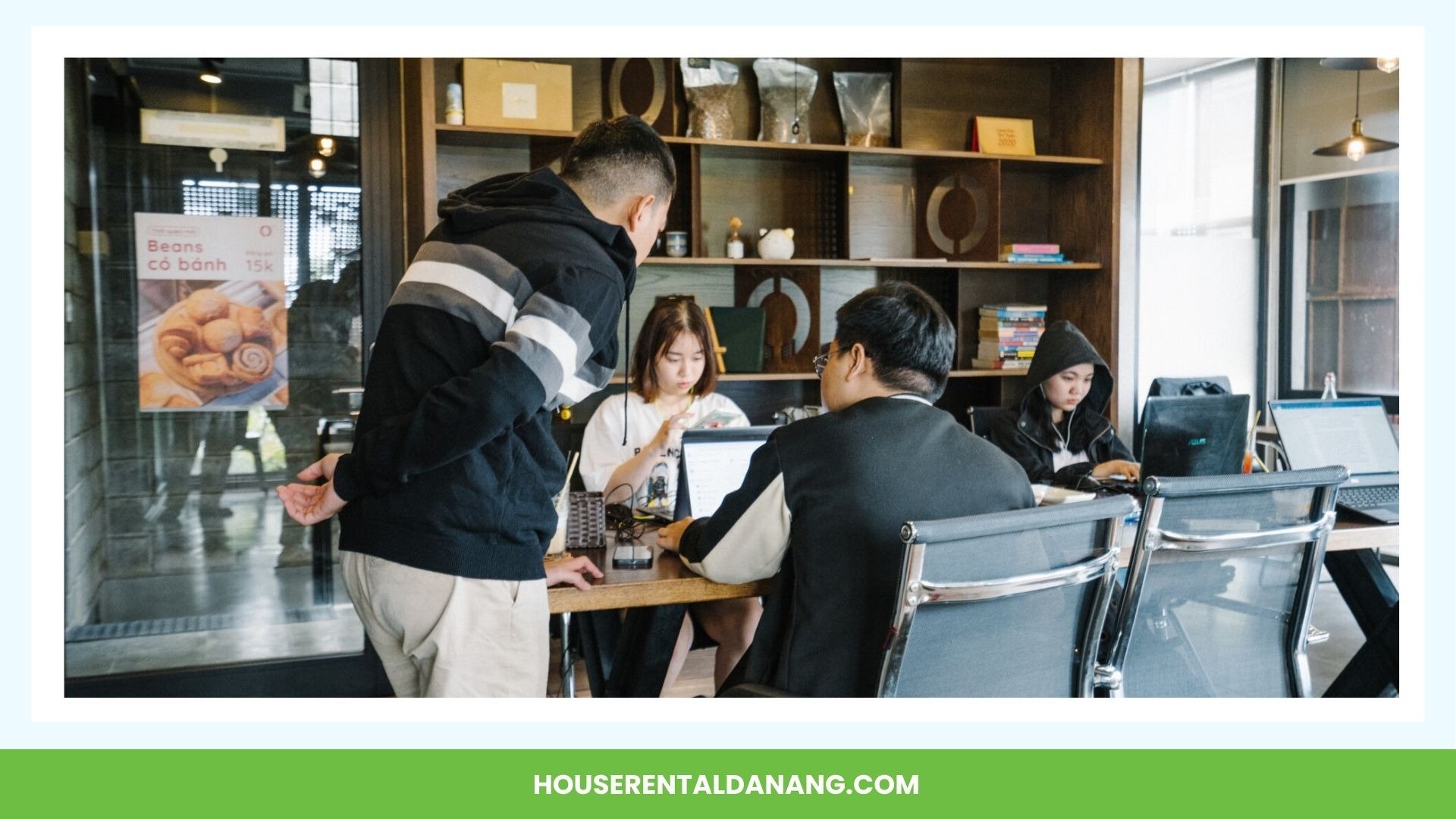 A group of four people are working on laptops at a table in a modern coworking space in Da Nang. A man is standing and leaning over to talk to the seated individuals, while shelves with books and decor serve as the background.