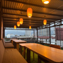 Rooftop seating area with wooden tables and cushioned benches under a corrugated metal roof. Hanging orange lights and a view of city buildings are visible.