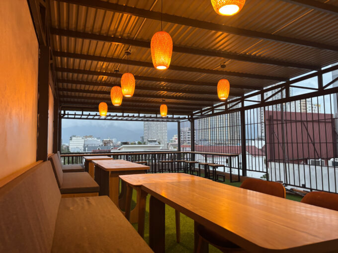 Rooftop seating area with wooden tables and cushioned benches under a corrugated metal roof. Hanging orange lights and a view of city buildings are visible.